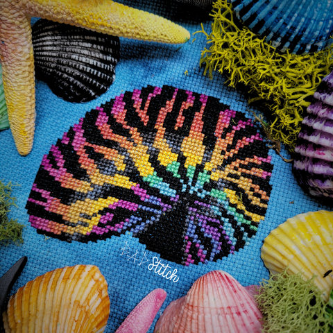 A cross stitch project on blue fabric, staged with green moss, painted seashells and starfish.  The cross stitch is of an empty nautilus shell with tiger stripe and rainbow colorings similar to the style of Lisa Frank. BAD Stitch.