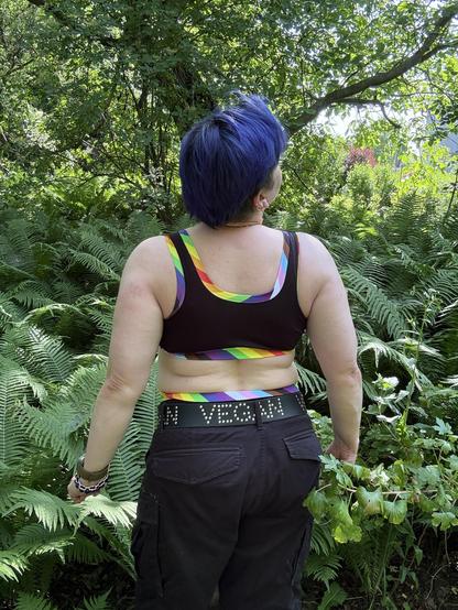 Osoba z krótkimi niebieskimi włosami na tle zieleni ogrodu (paprocie, drzewa) widoczna z tyłu, ubrana w czarny top/biustonosz z lamówkami w kolorach progress pride flag i czarne cargo szorty z paskiem z napisem VEGAN, znad których wystaje góra majtek w kolorach jak wykończenie biustonosza.
