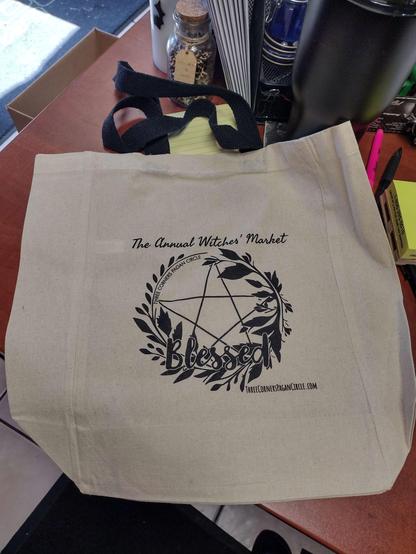 Natural colored bag with black handles. 
Print is a leafy circle with a pentagram inside and BLESSED written at the bottom of the circle. 

The Annual Witches' Market
ThreeCornersPaganCircle.com