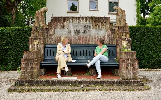 Photo d’un banc en bois peint en vert foncé inséré dans une structure massive en brique qui double à peu près sa hauteur et dont les côtés forment des gradins. Deux sculptures de perroquets viennent orner les deux angles supérieurs. Une inscription gravée dans une plaque en pierre mentionne le nom de Christine de Bosch Kemper, la date de sa naissance et celle de son décès. Deux femmes aux visages floutés prennent le café en discutant, assises sur le banc.