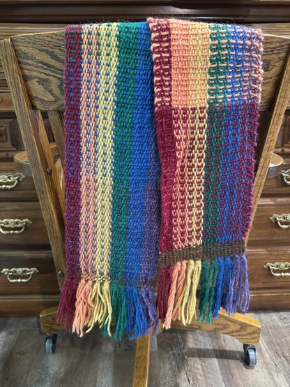 Over the back of a wooden chair at a wooden desk is draped a scarf that is bright and cheery. Along its length are colors of the rainbow. Across its width in blocks of color of rainbow from red to purple. One side the stripes of color along the length show strongly and the across show as little pops of color. The other side across shows more strongly almost making a checkerboard look. There is some brown woven in at both ends by the fringe representing juneteenth 6/19.