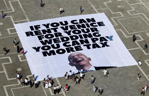 A large banner protest on a public square in Venice, Italy, staged in response to Jeff Bezos’ extravagant wedding. The banner features a massive portrait of Jeff Bezos laughing and the bold message: “IF YOU CAN RENT VENICE FOR YOUR WEDDING YOU CAN PAY MORE TAX.” Dozens of people surround the installation, taking photos and observing, highlighting the public discontent over wealth inequality and tax avoidance.
