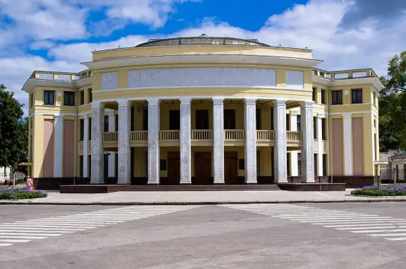 The image shows a large, classical-style building featuring a prominent façade with multiple columns. It has a yellow and white color scheme, and the architecture suggests it might be a theater or similar public venue. There is a clear blue sky with some clouds in the background, and the area around the building appears to be well-maintained, with flowers and a pedestrian crossing in front.

Image Credits: Wikimedia / Донор / CC BY-SA 3.0