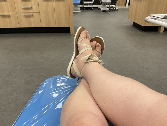 Emma zit ontspannen met haar benen gekruist tijdens het doneren van plasma bij de bloedbank. Ze draagt sandalen met een ivoorkleurig lint.