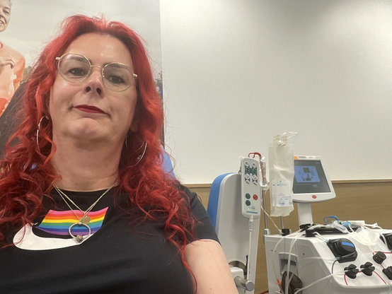 Emma, met rood haar en bril, glimlacht ontspannen tijdens haar plasmadonatie bij de bloedbank. Achter haar zijn medische apparaten te zien die gebruikt worden voor het afnemen van plasma. Ze draagt een zwart T-shirt met Nike logo en regenboogkleuren.