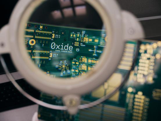 Magnifying glass examining an Oxide-branded circuit board, revealing the company logo and 'COSMO COMPUTE SLED' text on the green PCB with gold connectors