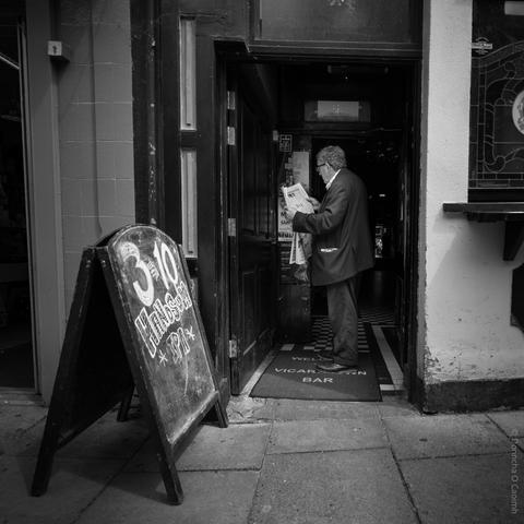 Black and white street photograph showing a man in a suit standing just inside the entrance of a pub, reading a newspaper. To the left, a chalkboard sign on the pavement advertises "3 for 10 Handsum IPA". The pub's doormat reads "WELCOME VICARSTOWN BAR". The scene captures a candid moment of daily life outside a traditional bar.