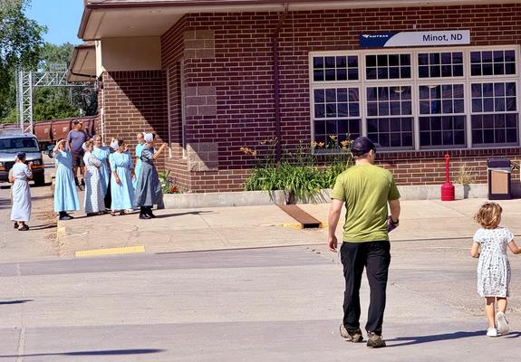 A group of women in blue dresses and bonnets stand outside a station in Minot, ND, while a man and a child walk away from the building. The scene features a brick building, greenery, and parked vehicles in the background.