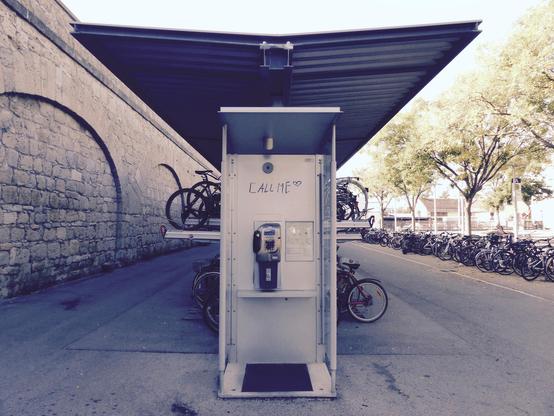 Eine öffentliche Telefonzelle an einem Bahnhof. Links davon ist die Bahnhofsmauer, dahinter und rechts von der Telefonzelle stehen viele Fahrräder. Die Telefonzelle hat keine Tür, und über dem Telefon selbst stehen in schwarzer Handschrift die Worte Call Me.