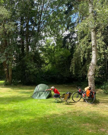 Zeltaufbau mit Mann, Abenteuerfahrrad und Campingstühlen unter hohen Bäumen