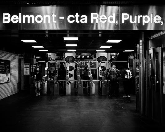The image shows the interior of the Belmont CTA station in Chicago. People are passing through turnstiles, and there's a large sign above indicating the red and purple lines. The space is busy, with bright overhead lights and dark flooring. The image is in black and white.