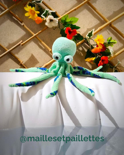 Poulpe au crochet vert menthe avec des tentacules à pointes bleu marine et vertes, posé au bord d'une piscine gonflable, sur fond de treillis en bambou décoré de fleurs colorées.