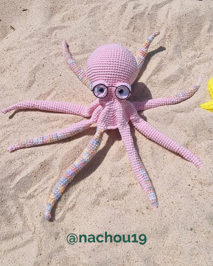 Poulpe rose crocheté, avec des lunettes rondes, posé sur du sable.