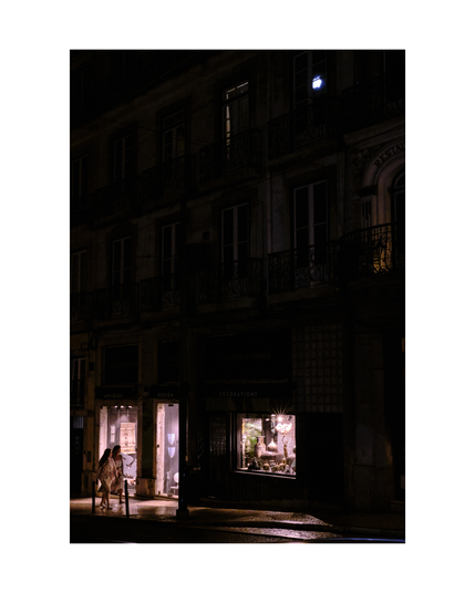building in lisbon by night with dark windows and out of shape moon reflection in an upper window with two women interestedly looking into and walking by the shop windows on ground floor