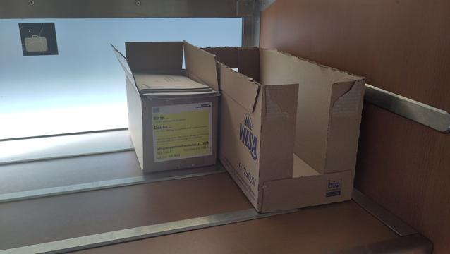 Zwei Kartonschachteln stehen in der Gepäckablage eines Zugs. Eine ist schon leer, sie enthielt 12 5 Deziliter PET Wasserflaschen. Die zweite enthält Umschläge, sie ist mit der Aufschrift beschriftet "Fahrgastrechte-Formular F 2023, 200 Stück, Version 05.2018, Karton 05.952"