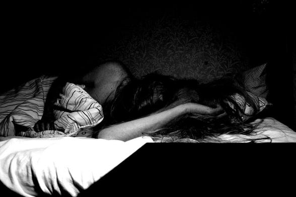 A high contrast image in black and white of a person lying in bed. The only thing visible of the person is a shoulder, two arms, and a head bent down so non of the face can be seen behind all the hair that is spread out all over the pillow. One arm infront of the head and rhe other one is resting upon the quilt. At the low right front, part of a dark table is seen and to the low left some bright over-exposed sheet. In the background, some of the light expose some plant pattern on the wall behind the scene.
