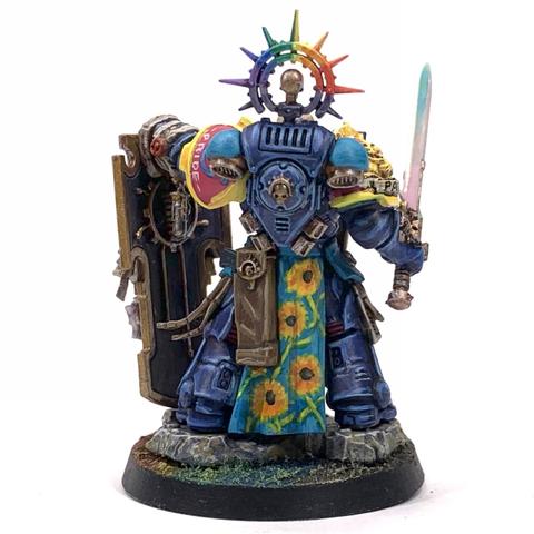 Large warrior in dark blue armor with pansexual color (blue, yellow, and magenta) decorations. He carries a pearlescent sword and a large bulwark shield with rainbow colors behind a skeleton holding an hourglass and wrapped in scrolls that say "Pride". His tabard has sunflowers over light blue. He has a rainbow halo over his bear head, which stares out beyond the shield in defiance.
