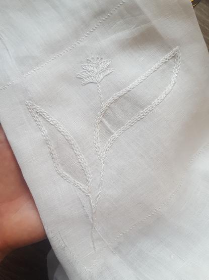 A simple flower with two large leaves embroidered in white onto white linen fabric. The sleeve seam runs under one of the big leaves, and there's only one layer of fabric on the leaf part, and two elsewhere on the patch. The cut out top layer has been affixed with two lines of chain stitch, making for a sort of lily of the valley type leaf pattern, and a little flower has been added with buttonhole stitch, quite abstracted. The patch has been sewn on from the reverse, showing slanted lines of whipstitch through. It's all slightly shiny, being mercerised cotton embroidery thread.