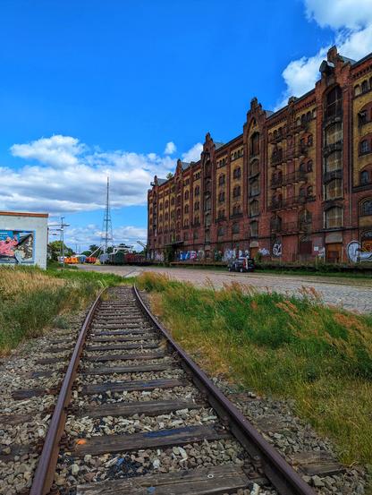 A low-angle, wide shot shows a single railway track curving slightly to the left in the foreground, with loose ballast and weeds growing between the sleepers. To the right of the track, tall green and golden grasses line a wide, unpaved area. Dominating the right side of the frame is a long, multi-storey brick building with numerous windows, displaying signs of age and some graffiti at its base. Its industrial architecture is prominent under a bright blue sky with scattered white clouds. In the distance, a tall, slender antenna tower is visible, and further down the track, a white building with graffiti and other industrial elements can be seen. The scene evokes an abandoned or disused industrial area.