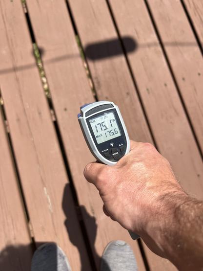 A hand holds a digital infrared thermometer displaying a temperature of 175.1°F. The background features a wooden deck with visible grooves.