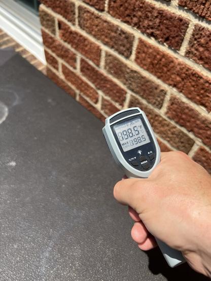 An infrared thermometer displays a temperature reading of 198.5°F from a dark brown deck box. It is held in a hand, with a brick wall partially visible in the background.
