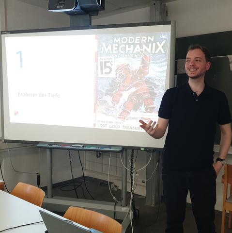 Der Referent vor einer Folie mit dem Titel "Eroberer der Tiefe" und darauf das Cover einer historischen Ausgabe der "Modern Mechanix" mit futuristisch wirkendem Tiefseetauchanzug darauf.