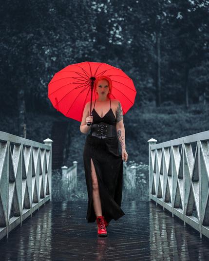 Woman with red hair and tattoos walks down a wet wooden bridge holding a red umbrella. She is wearing a long black dress with a slit and red boots. The background features a blurred view of trees in a rainy, overcast setting.