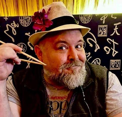 Head-and-shoulders color photo of writer Frank Oreto, a man with a trimmed, graying beard and mustache. He wears a dark gray fleece vest over a gray tee, accessorized with glasses on a chain and a small-brimmed straw hat accentuated with a black band and a magenta orchid. He sits in what looks to be a restaurant booth and wields chopsticks with an eager expression.