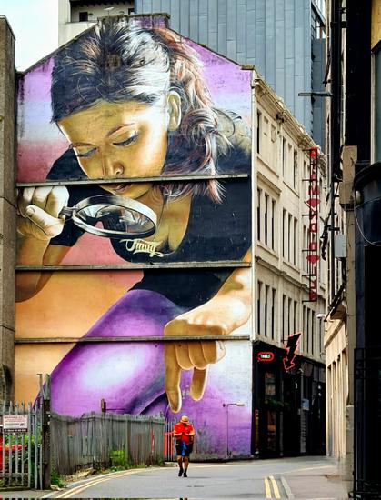 A mural of a young woman with a magnifying glass who appears to be plucking a passing pedestrian from the street below.