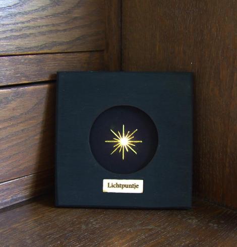 A Lichtpuntje artwork stands in a quiet, dark corner of a wooden work desk. A small light glows brightly at the center. The piece is framed in a handmade black wooden frame and features a label that reads "Lichtpuntje."