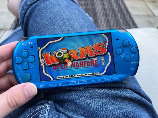 A photo of my blue Sony PSP resting on my thigh. The screen it’s showing the titles for the game Worms: Open Warfare.