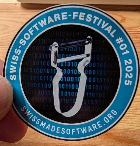 Photo of a "Swiss-Software-Festival" sticker with a blue border, and a silver potato peeler in the centre on dark background.