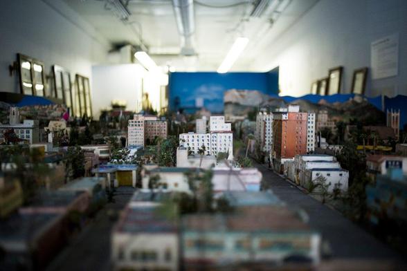 The image depicts a detailed model of a cityscape, likely part of an exhibition or display. The model includes a variety of buildings, ranging from low-rise structures to taller apartment buildings, with a mix of colors including white, orange, and blue. The buildings are arranged along streets, with some greenery interspersed among them, suggesting a realistic urban environment. The background features a painted mural of a mountainous landscape, adding depth to the scene. The setting appears to be indoors, with fluorescent lighting illuminating the model. The walls are adorned with framed pictures, and there is a sign on the right side, possibly providing information about the model. The overall composition is intricate, with a focus on the detailed architecture and layout of the city.

Provided by @altbot, generated privately and locally using Ovis2-8B