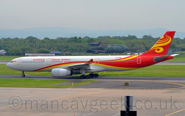 Side view of a twin engined jet airliner taxiing from right to left.
The plane is mostly white, with a red and yellow stripe swooping from under the lower forward fuselage and over the wing, with the yellow descending back under the rear fuselage, while the red takes over the rear fuselage and tail, with additional yellow swirls at the top and on the tail.
There are red "Hainan Airlines" titles on the upper forward fuselage, in a Chinese script forward if the mid-cabin door, and in English aft of that door.
The black registration "B-304K" is on the upper rear fuselage.
Grey concrete apron and black tarmac taxiway fill the foreground, with trees stretching across the background under hazy grey sky.
In the middle of the background, a green fire training rig, vaguely resembling a 3-engined Boeing 747, is sitting there looking a bit sorry for itself.