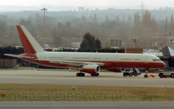 Side view of a twin engined jet airliner parked facing to the right.
The plane has white upper surfaces and red lower surfaces, with the tail being white at the front and red at the rear.
There is a grey and dark blue tug attached to the planes nose-wheel by a long red pole.
Green grass stretches across the foreground, separating the runway from a taxiway, with trees and buildings stretching into the distance in the background, slowly vanishing into hazy skies.