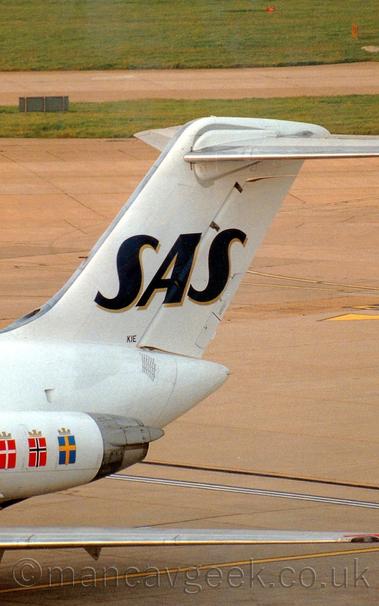 Close-up of the white rear fuselage and tail of a twin engined jet airliner with the engines mounted on the sides of the rear fuselage.
The plane is parked facing to the bottom left of the frame.
There are 3 flags on the engine pods, on the left is a red flag with a white cross, in the middle is a red flag with a black cross that is outlined in white, and on the right is a blue flag with a yellow cross.
Each flag has a gold crown on the top.
The tail is white, with large black letters "SAS" with a gold drop-shadow, with very small letters "KIE" at the base of the tail.
Grey concrete apron fills the background, with a large grass area at the top of the frame.