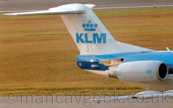 Close-up of the white tail of a twin engined blue and white jet airliner taxiing from left to right.
The planes white engines are mounted on the sides of the rear fuselage, and have small "Fokker F100" text at the front.
The planes tail is white, with large blue letters "KLM" under a stylised blue crown in the middle.
Yellowing grass fills the background, with a black taxiway stretching across the top of the frame.