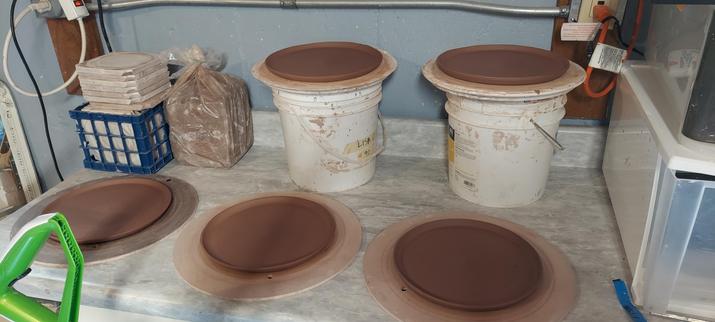 5 of 8 red/brown stoneware plates drying on a countertop. These are hybrid plates. They start as 1/4" slabs that I transfer onto bats, then trim to a 10.5" circle. They are compressed on the wheel and the edge is lifted to form the plate rim.