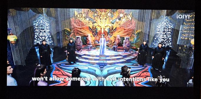 Bai Shuo standing in front of the throne in Bright Moon Palace. The floor before her is a sea of swirling shapes in blue, turquoise and red. The wall behind her is decorated with a gigantic stylished flower pattern, that explodes from behind the throne in yellow and orange.