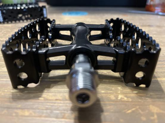 Das Bild zeigt eine Nahaufnahme eines schwarzen Fahrradpedals mit einem einzigartigen Design mit mehreren Löchern und gezackten Kanten. Es ist auf einer Holzoberfläche positioniert und hebt seine detaillierte Struktur und die Gewindeverbindung auf einer Seite hervor.