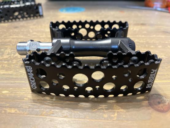 Ein Nahaufnahmebild eines schwarzen Fahrradpedals mit einer strukturierten Oberfläche für den Griff. Das Pedal verfügt über mehrere Löcher und eine Gewindeachse zur Befestigung. Der Hintergrund ist eine Holzoberfläche mit Hinweisen auf Werkzeuge und andere Fahrradkomponenten.