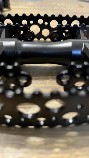 Das Bild zeigt eine Nahaufnahme von schwarzen Fahrradpedalen, die ein detailliertes Design mit Löchern und Graten zeigen. Der Hintergrund ist verschwommenes Holz, das die Pedale betont.