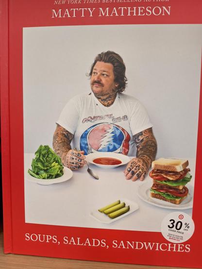 Cover of Matty Matheson's book "Soups, Salads, Sandwiches." He has a bowl of what looks like tomato soup in front of him, a huge sandwich to his left, and a salad to his right. He's wearing a "Steal Your Face" Grateful Dead T-shirt.