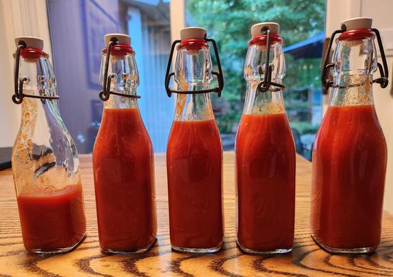 5 glass bottles on a wooden table, each capped and closed.  Four on the right are full, the one on the left is only about 1/3 full. They contain bright red hot sauce.