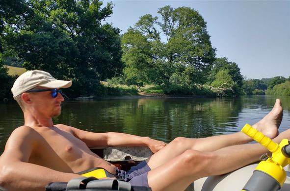 A white man (me) wearing sunglasses, a pale cap and navy shorts is sat with his feet up in an inflatable boat. It's sunny, and behind the man we see the River Dart, lined with trees and looking beautiful. The sky is pale blue and cloudless.