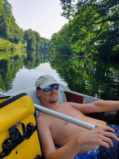 A white man (me) wearing sunglasses, a pale cap and navy shorts is sat with his feet up in an inflatable boat. He's holding an oar. Beside him there's a yellow bag.
It's sunny, and behind the man we see the River Dart, lined with trees and looking beautiful. The sky is pale blue and cloudless.