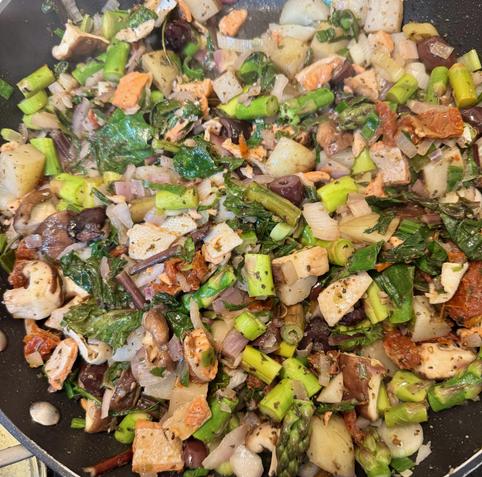 a skillet with a mix of chopped, cooked foods: asparagus, potatoes, mushrooms, onion, and dandelion greens, with small pieces of salmon also visible.