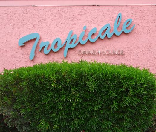 "Tropicale" sign in turquoise script, tilted at angle from low to high, left to right, against a bright pink stucco wall. Below the sign is a bright green trimmed hedge.