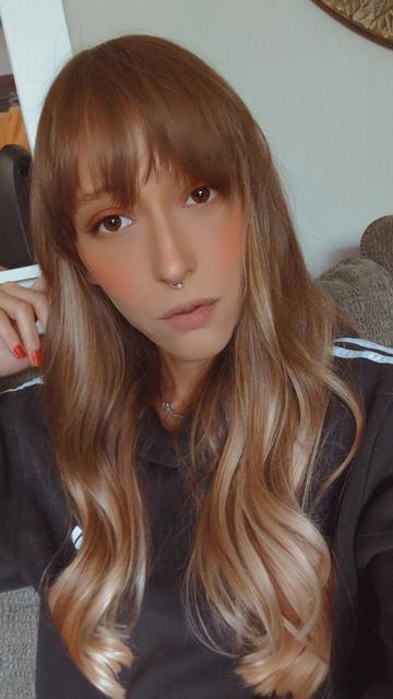 Image of Scarlet sitting on a couch. She has long, wavy, light brown hair with bangs. She's wearing a black top with white stripes down the arms. She has a subtle makeup look with rosy cheeks, a natural lip color, and a septum piercing. Her expression is calm and thoughtful.