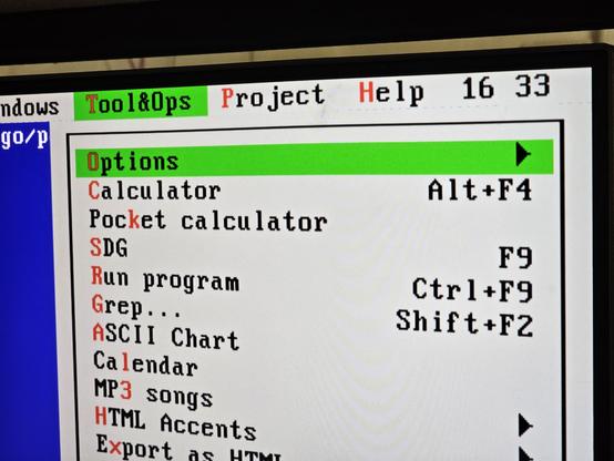 Photo of a right corner of a computer screen.

Grey menubar with green highlight is shown in 80×25 text mode in the typical VGA font. Background of the “window” is blue with white text, like usual in many DOS editors.

The menubar ends with items “Windows”, “Tool&Ops”, “Project” and “Help”. Next to the right-most item (Help), current time (16:33, with the colon blinking – hidden in the photo) is displayed. The menus have their first character highlighted in red, as a shortcut with the Alt key.

The “Tool&Ops” menu is open. Menu items:

Options (submenu arrow is shown)
Calculator (Alt+F4)
Pocket calculator
SDG (F9)
Run program (Ctrl+F9)
Grep... (Shift+F2)
ASCII Chart
Calendar
MP3 songs (submenu)
HTML Accents (submenu)
Export as HTML (submenu)

The rest of the menu is cropped out of the photo.